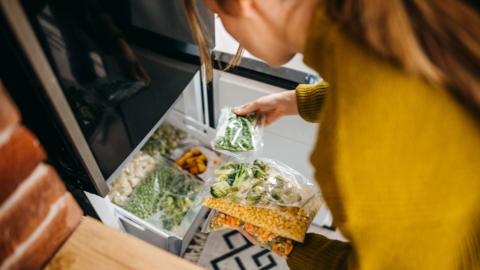 Woman putting container with frozen mixed vegetables to refrigerator