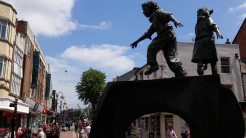 Large black statue in the foreground of a Cobbler's last with a boy and girl playing on it. A town centre shopping street is visible behind the statue.