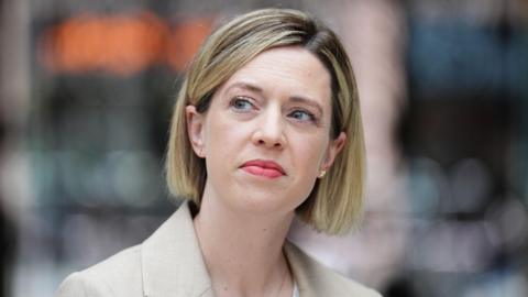 A woman with a blonde bob, wearing a beige blazer and a white top staring off into the distance.