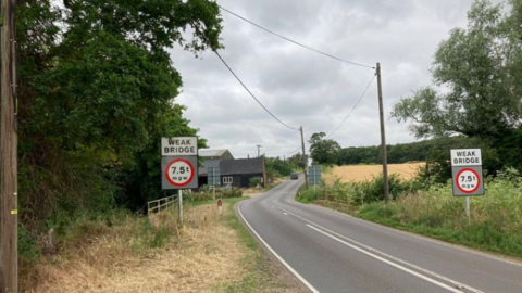 A general view of Bridge Farm Bridge at Stowlangtoft near Bury St Edmunds in Suffolk. A small bridge can be seen with signs that read "weak bridge" and the weight limit. A building can be seen in the background and there are several pylons and overhead wires that run along the length of the road. A car can be seen on the road in the distance. There are trees and fields on either side of the road.