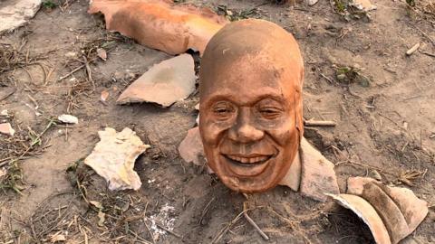 The severed head of from the statue of Nana Akufo-Addo, bronze in colour, lying on the ground. There are fragments of other parts of the statue on the dirt ground