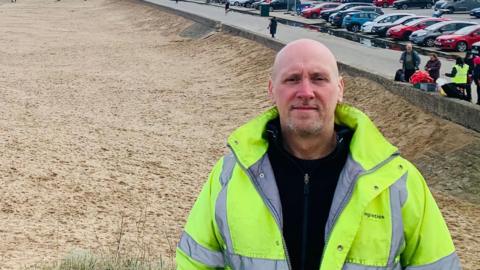 Carl Quartermain is standing on a sandy beach and looking directly at the camera. He is bald, has a grey beard and is wearing a yellow high-vis jacket over a black fleece. On the land, there are dozens of cars in the car park and rows of houses further along the shore.