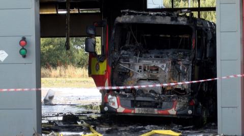 The front of a fire engine has completely melted away with the remains of a gutted appliance, within a fire station with debris lying on the floor and a emergency tape restricting entry.