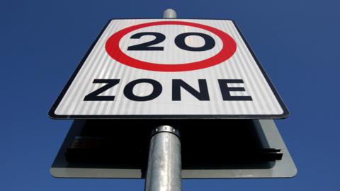 A white signboard with text saying 20 in black, surrounded by a red circle and the word Zone below it