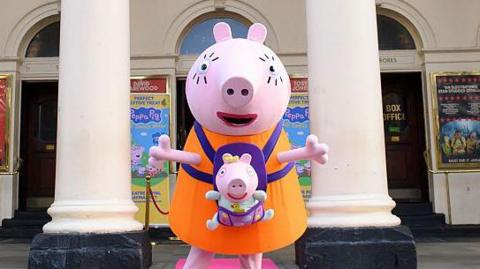 A person in a large pig costume stands outside a theatre with their arms spread wide. A smaller pig in a baby carrier is attached to their chest. The theatre is visible in the background with production posters on the walls.