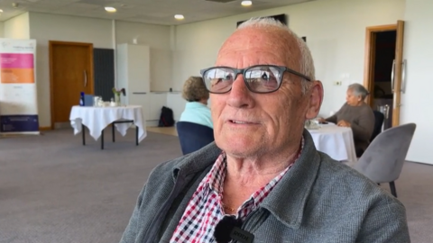 An elderly man wearing black-rimmed glasses and a checked shirt, sitting down 
