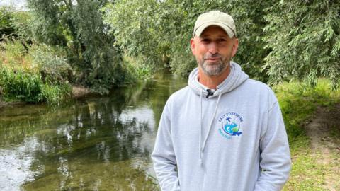 A man wearing a grey hoodie and baseball cap smiles as he stands on a grassy bank in front of a clear chalk stream with green trees behind him.