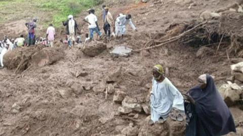 A group of people can be seen on a muddy hillside looking at the wreckage caused by the landslide.