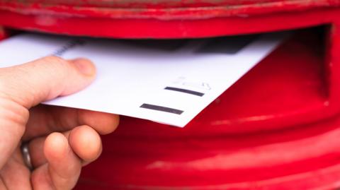 A hand holding a letter and posting it in a red post box.