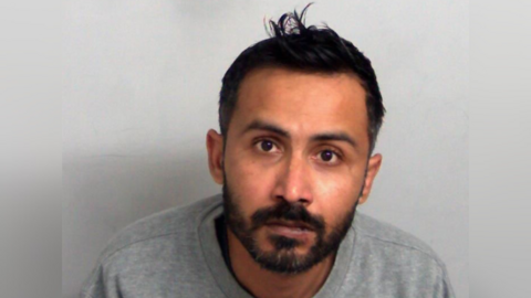 A custody photo of Naeem Shahani, who is wearing a grey sweater and looking at the camera looking worried. He has black hair which is slightly spiked up in the middle.