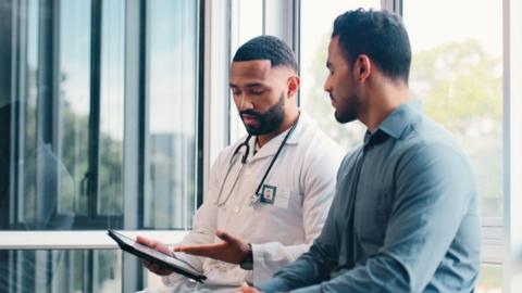 A male doctor points at a clipboard with his hand whilst showing it to a young male patient.