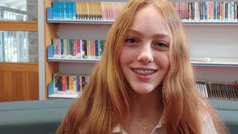 A teenage girl with long thick blonde hair over her shoulders. She is wearing a white shirt, smiling, and sitting on a green sofa. She is sitting in a library and behind her are shelves and books. 