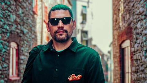 DJ Ridney wearing sunglasses and a black shirt standing in a narrow street
