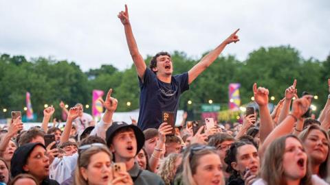 Festivalgoers enjoy a performance during BBC Radio 1's Big Weekend. There are fans in the crowd who are singing along and one who is raised up with his arms in the air. 