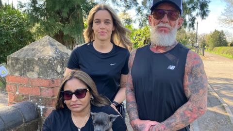 A group of three people stand outdoors near a brick wall, with one person sitting and holding a small dog.
