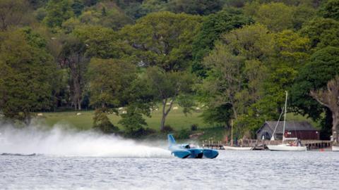 Bluebird K7 "planes" on Coniston Water. The blue craft is throwing up a large spray behind it as it glides on top of the lake's surface. On the far shore, sheep are grazing on an incline.