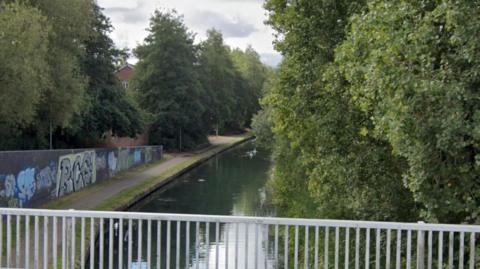 A Google Maps image of a canal. We can see a metal gate at the bottom of the screen. Then the canal bends off to the right with some birds swimming in it. There is a wall along the canal with graffiti on it. To the right of the screen there are trees and bushes. 
