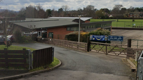 A driveway leads towards a school building, with playing fields in the background.