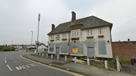 The pub sits on a right hand bend away from houses or other properties. It is painted cream but has metal covers on windows and the pub's name can be seen in black letters above the former entrance. It has two levels with uncovered windows higher up.