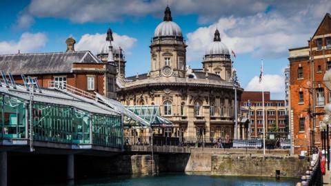 A waterfront scene with a glass walkway on the left and buildings in the distance.