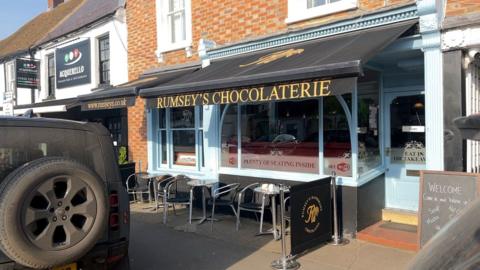 The front of Rumsey's Chocoloterie in Thame. The black awning displays the shop's name, and several tables and chairs are seen out the front. The building is a Georgian red brick terrace, with a light blue front door.