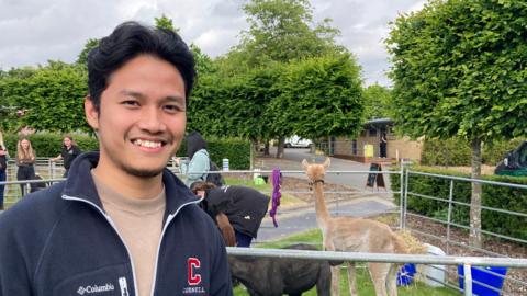 A man with a navy blue zip up top and camel-coloured top beneath is standing in front of a pen of llamas and other animals in pens. Blue feeding buckets can be seen to the side of the closest pen. There is greenery and trees in the background.