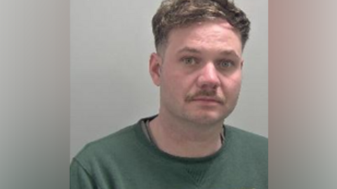 A police mugshot of a man with short brown hair, a moustache and a green jumper.