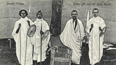 A historic black and white photograph of four men in white robes holding spears and shields