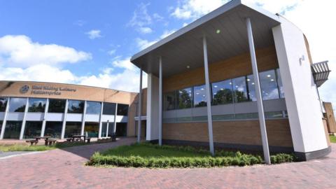 A modern building which is white with sandy brickwork has windows going across the middle and a roof coming across the top with white poles going down into a triangular grass verge in front of it. Next to it is more of the building with benches placed outside and the words 'East Riding Leisure Haltemprice' on the building in grey lettering.