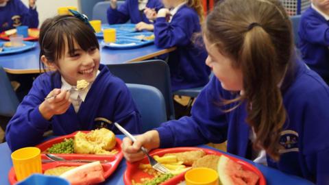 Two school children are eating from their dinner trays during lunchtime in their school canteen. Both are girls with brown hair wearing blue school cardigans over white shirts. One has a jacket potato and peas on her tray and both have a big slice of watermelon for dessert.