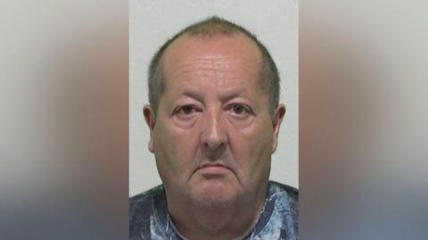 Mugshot of John Briggs. He has very short balding dark hair, is clean shaven with a large head and downturned mouth. He is looking very morose.
