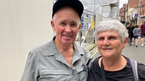 An older man and woman look at the camera. The man is wearing a cap and has a couple of shirt buttons un-done as it is a warm day. The woman is wearing a rucksack and a black T-shirt. Behind them is scaffolding near York Minster.
