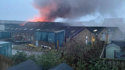 Image of the back of what looks like a farming estate, with black smoke billowing above the buildings. Just above some of the far-away buildings, the smoke is orange.