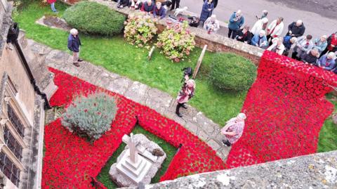 Looking down from church bell tower onto the poppy cascade, lots of red knitted flowers stitched closely together with people coming to look over the wall, with the war memorial in the middle