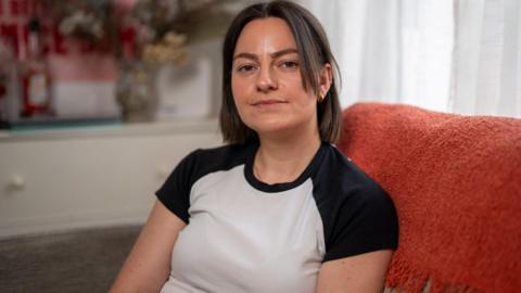 Lucy Asson has shoulder-length dark hair and is wearing a white t-shirt with black shoulders