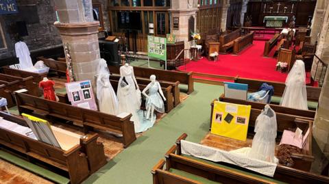 A church interior, seen from a height, with six standing mannequins clothed in long white wedding dresses, three with veils, and one seated one displayed between the pews. A smaller mannequin wears a long red dress, and there are a number of poster boards with photos.
