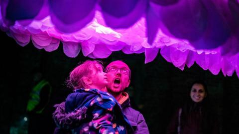 A man golds a young girl under a purple illuminated installation that looks like petals on the bottom of a huge flower. It is dark and their faces are lit up purple