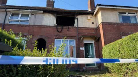 Police crime scene tape across the front of a residential property, which has its window burnt out.