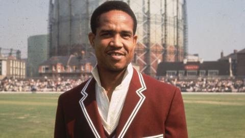 Gary Sobers wears a red suit beside a cricket ground in an image from 1966