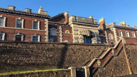 The exterior of HMP Exeter. It is a historic looking building of red brick with ornate granite block work and many chimneys. There is an inscription engraved in the stone above the entrance which reads: DEVON COUNTY PRISON. In the foreground is a stone wall with steps running through it.