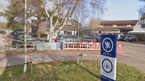 A general view of the exterior of the Brookes UK School. A car park can be seen outside the school with cars parked in it. Large trees are in front of the main building.