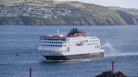 The Manxman ferry, which is back, white and red, arriving in a sunny Douglas Bay with the coastline of Onchan Head in the background.