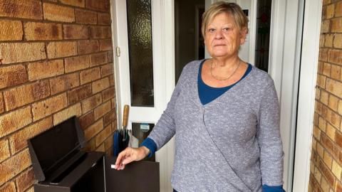 Jen Caple, an older woman with short blonde hair, stands in front of her white front door, holding open the door of her empty brown post box. She is straight faced and is wearing a marl grey wrap cardigan with a long-sleeved blue top beneath it and a gold chain necklace. There is an umbrella propped up against the window pane beside her front door.