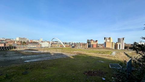 A view of the grassy field where the arena is supposed to be built. The white, curved Millennium bridge is poking out from over the hill along with the red and yellow Baltic art gallery.
