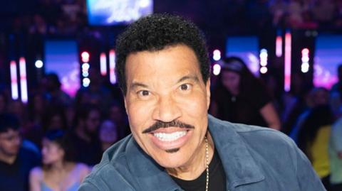 A black man with short, curly, black hair and facial hair is smiling toward the camera. He is wearing a a blue suede jacket with a black t-shirt underneath and a silver chain around his neck. 