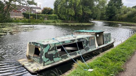 Rusted empty canal boat sat on the side of a canal tethered to a post in the grass 