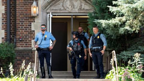 Four police officers, three with bulletproof vests and one with a helmet and gun standing outside an open entrance. There are steps leading to the doorway