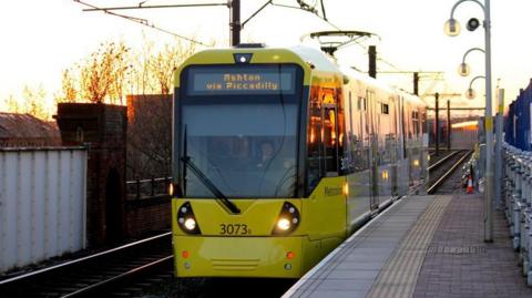 A yellow and grey tram on a track at a platform outside. The sun is setting in the background. 