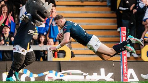 Leeds Rhinos player Harry Newman, in dark blue shirt and white short, dives over the try line to score against Huddersfield Giants while to his left the Leeds mascot Ronnie the Rhino raises his arms in celebration and fans applaud in the background.