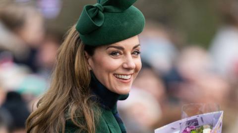 Kate is smiling and wearing dark green and a matching green hat. She has a green and navy checkered scarf around her neck.
